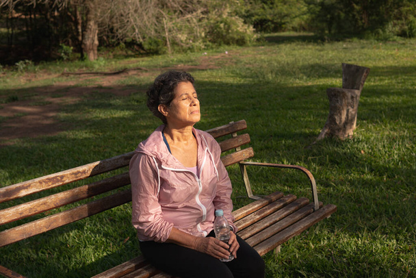Preciosa brasileña jubilada sentada en el banco, sosteniendo una botella de agua y disfrutando del sol. Al aire libre en zona verde. Sed, hidratación, alegría, concepto de vitalidad. - Foto, imagen