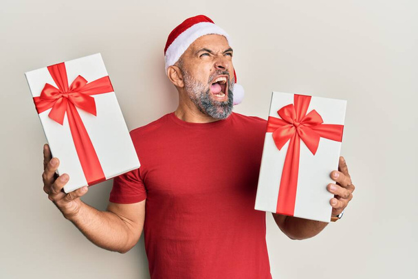Мужчина средних лет в рождественской шляпе и с подарками, злой и безумный, кричащий расстроенный и разъяренный, кричащий от злости. гнев и агрессивная концепция.  - Фото, изображение