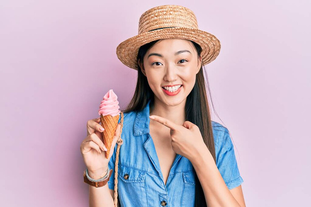 Young chinese woman wearing summer hat holding ice cream smiling happy pointing with hand and finger  - Zdjęcie, obraz