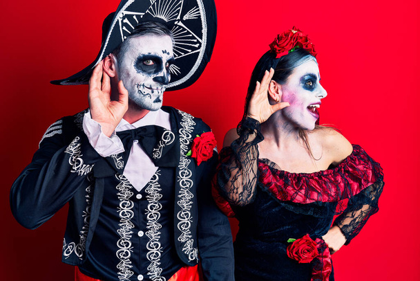 Junges Paar im mexikanischen Totenkostüm über rotem Lächeln mit der Hand über dem Ohr, das Gerüchten oder Klatsch lauscht. Konzept für Gehörlosigkeit.  - Foto, Bild