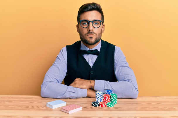 Hezký hispánský krupiér sedící na stole s pokerovými žetony a kartami s vážným výrazem na tváři. jednoduchý a přirozený pohled do kamery.  - Fotografie, Obrázek
