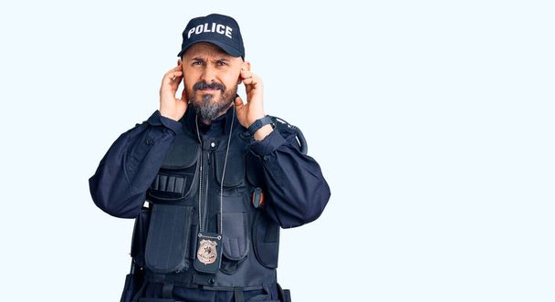 大音量の音楽のノイズのためにいらいらする表情で指で耳を覆う警察の制服を着た若いハンサムな男。聴覚障害の概念.  - 写真・画像