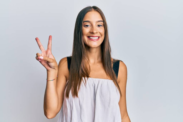 勝利サインをしているカメラには幸せそうな顔をして笑顔でカジュアルな服を着ている若いヒスパニック系の女性。2番.  - 写真・画像