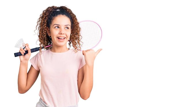 Beautiful kid girl with curly hair holding badminton racket and shuttlecock pointing thumb up to the side smiling happy with open mouth  - Photo, Image