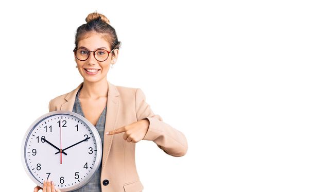 Beautiful caucasian woman with blonde hair wearing business jacket and holding clock smiling happy pointing with hand and finger  - Photo, image