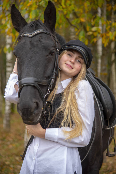  portrait of a girl with blonde hair , wearing a cap, and a Bay horse on the background of a forest.  - Foto, imagen