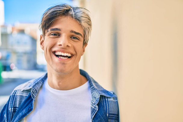 Νεαρός Ισπανός χαμογελά χαρούμενος ακουμπώντας στον τοίχο της πόλης. - Φωτογραφία, εικόνα