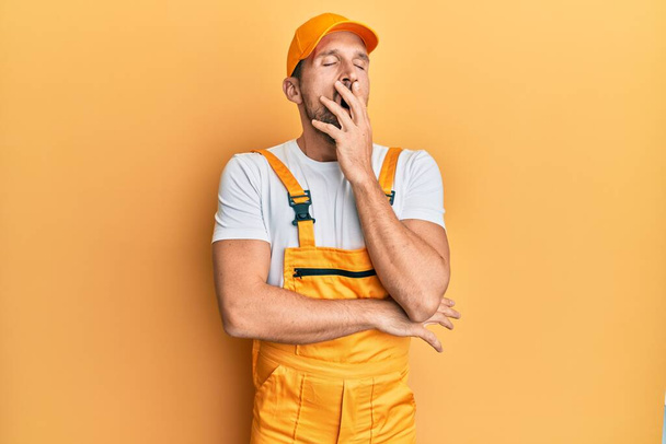 Young handsome man wearing handyman uniform over yellow background bored yawning tired covering mouth with hand. restless and sleepiness.  - Photo, Image