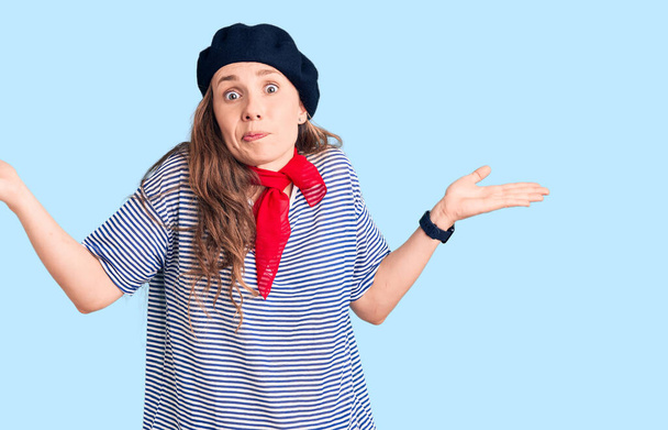 Young beautiful blonde woman wearing french beret and striped t-shirt clueless and confused expression with arms and hands raised. doubt concept.  - Photo, Image