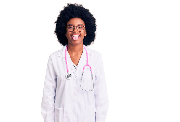 A fiatal afro-amerikai nő orvosi kabátot és sztetoszkópot visel, és vicces arckifejezéssel kidugja a nyelvét. érzelem koncepció.  - Fotó, kép