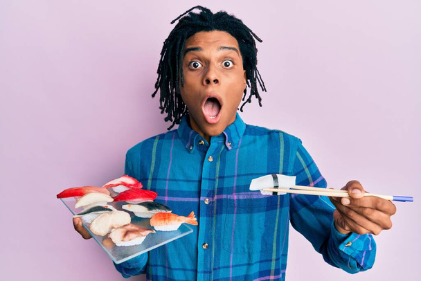 Young african american man eating sushi using chopsticks afraid and shocked with surprise and amazed expression, fear and excited face.  - Фото, изображение