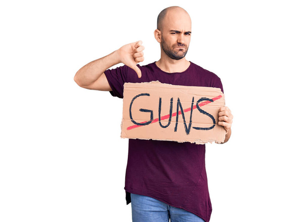Young handsome man holding prohibited guns banner with angry face, negative sign showing dislike with thumbs down, rejection concept  - Photo, Image