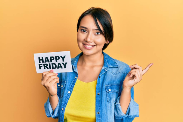 Piękna młoda kobieta z krótkimi włosami trzymając szczęśliwy piątek wiadomość papier uśmiechnięty szczęśliwy wskazując z ręki i palca na bok  - Zdjęcie, obraz