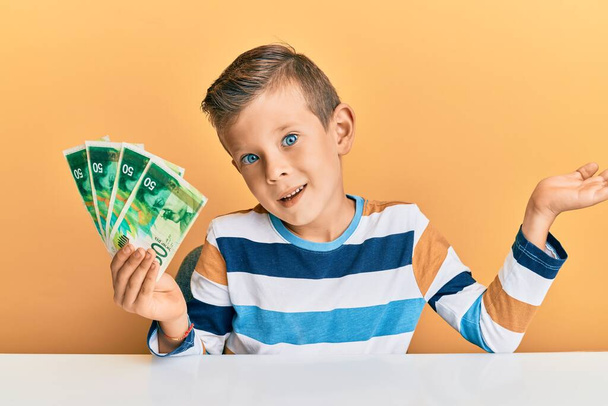 Schattige blanke jongen met israël 50 shekels zittend op de tafel vieren prestatie met een vrolijke glimlach en winnaar uitdrukking met opgeheven hand  - Foto, afbeelding
