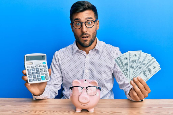 Hezký hispánec drží dolary počítání úspor v šoku tvář, vypadá skeptický a sarkastický, překvapený s otevřenými ústy  - Fotografie, Obrázek