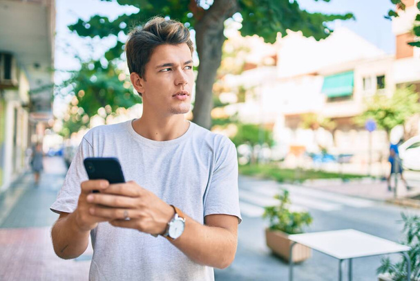Młody biały mężczyzna z poważnym wyrazem twarzy przy użyciu smartfona w mieście. - Zdjęcie, obraz