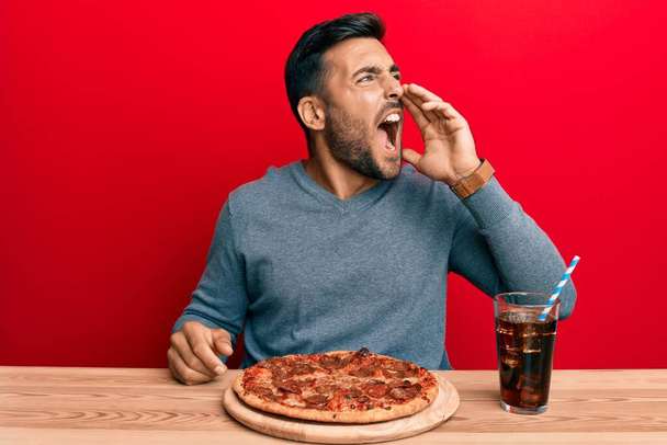 Όμορφος Ισπανός που τρώει νόστιμη πίτσα πεπερόνι φωνάζοντας και ουρλιάζοντας δυνατά στο πλάι με το χέρι στο στόμα. έννοια επικοινωνίας.  - Φωτογραφία, εικόνα