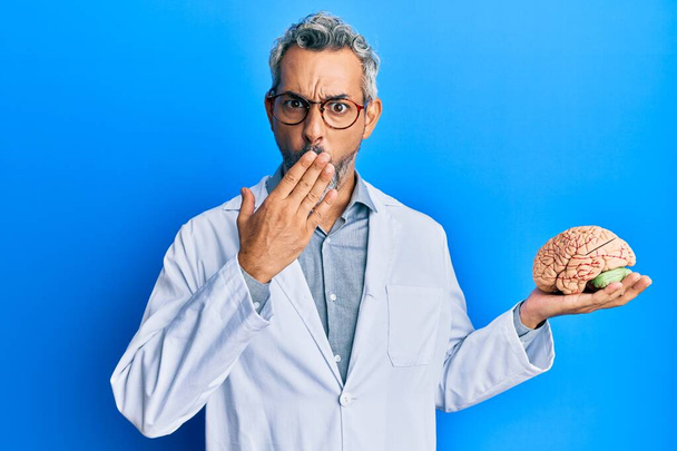 Siwy mężczyzna w średnim wieku, noszący płaszcz lekarza, trzymający mózg zakrywający usta dłonią, zszokowany i bojący się pomyłki. zaskoczona ekspresja  - Zdjęcie, obraz