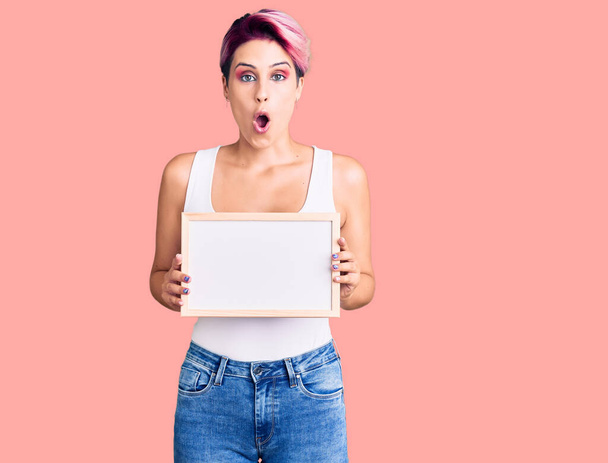 Young beautiful woman with pink hair holding empty white chalkboard scared and amazed with open mouth for surprise, disbelief face  - Photo, Image