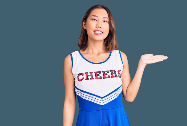 Young beautiful chinese girl wearing cheerleader uniform smiling cheerful presenting and pointing with palm of hand looking at the camera.  - Photo, Image