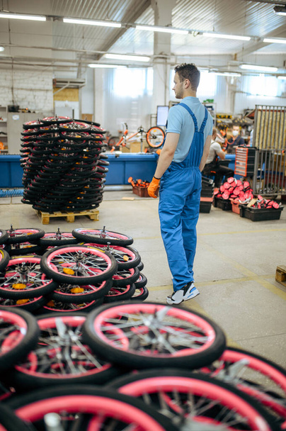 Велосипедна фабрика, працівник у стосі велосипедних коліс. Чоловіча механіка в уніформі встановлює частини циклу, складальна лінія в майстерні, промислове виробництво
 - Фото, зображення