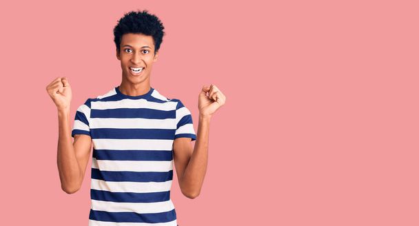 Gündelik giysiler giyen genç Afro-Amerikalı adam kollarını kaldırıp, gözleri açık bir şekilde başarıyı kutluyor ve şaşırıyor. Kazanan konsept.  - Fotoğraf, Görsel