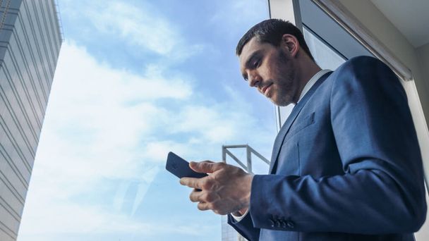Low Angle Shot of Successful Businessman in a Suit Standing in His Office, Using Mobile Phone while Standing next to Window with Sky View in Big City. - Fotoğraf, Görsel