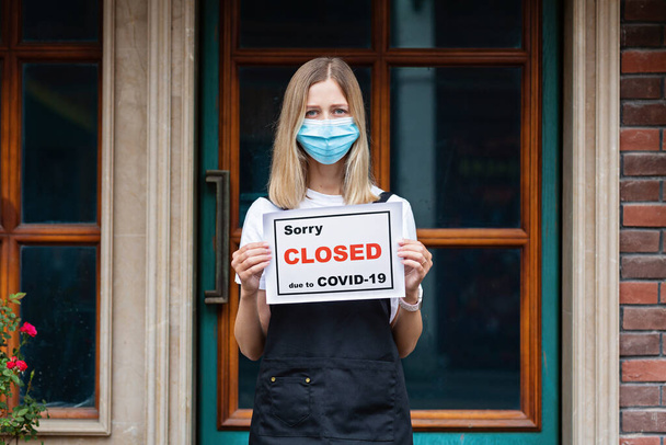 Caucasian waitress woman wearing medical mask holds SORRY WERE CLOSED. Coronavirus pandemic. Government shutdown of restaurants, shopping stores, non essential services - Fotó, kép