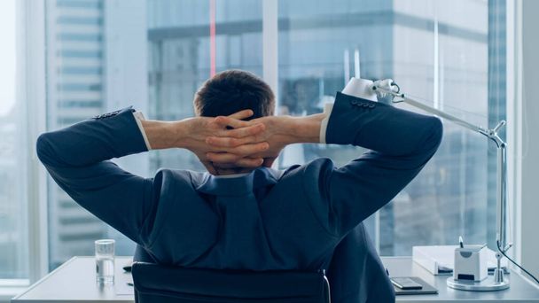 Back View of Confident Businessman Sitting at His Desk and Stretches. Hes Fatigued and so Tired. Successful Corporate Man Doing Important Job. Modern Office Environment Has Window with Big City - Foto, Imagen