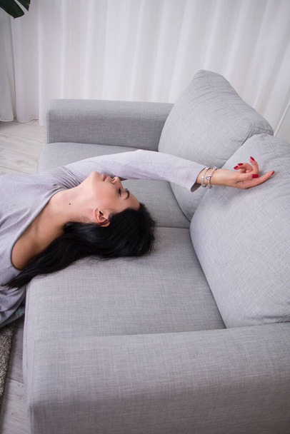 Chica en un suéter gris se sienta en el suelo cerca de un sofá gris - Foto, imagen