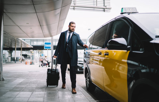 Handsome male passenger in formal outfit sitting in yellow cab getting to business meeting during travel trip, Caucasian man lawyer with baggage suitcase ordering taxi transport at airport exterior - Photo, Image