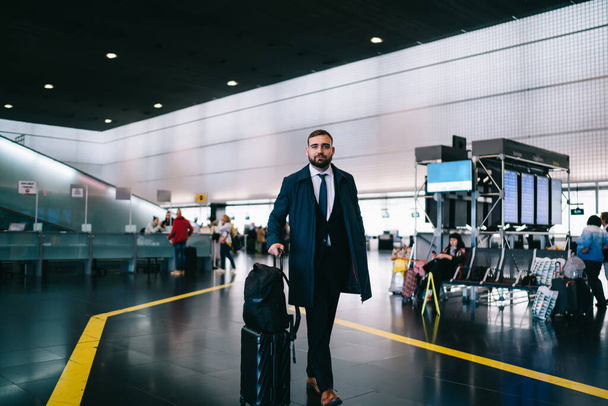 Full length portrait of formally dressed corporate director with luggage suitcase and travel backpack walking inside of airport and looking at camera, proud CEO posing during business trip - Фото, изображение