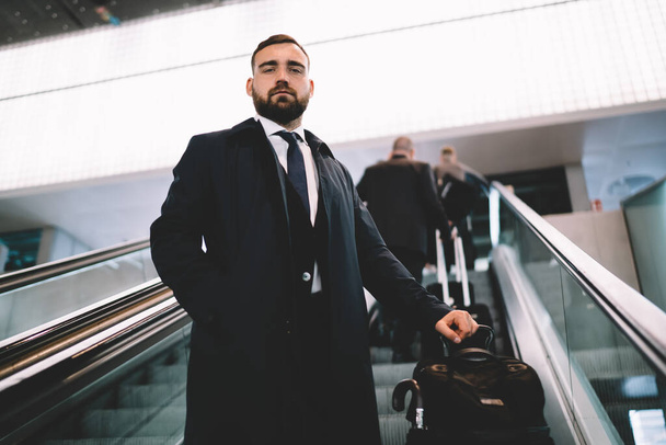 Half length portrait of handsome male commuter with baggage luggage looking at camera while getting up on electric stairs, Caucasian businessman in formal clothing have work trip from airport - Foto, Imagen