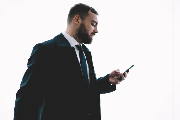 Side view of bearded successful guy in formal suit using smartphone and looking at screen while standing against white background - Zdjęcie, obraz