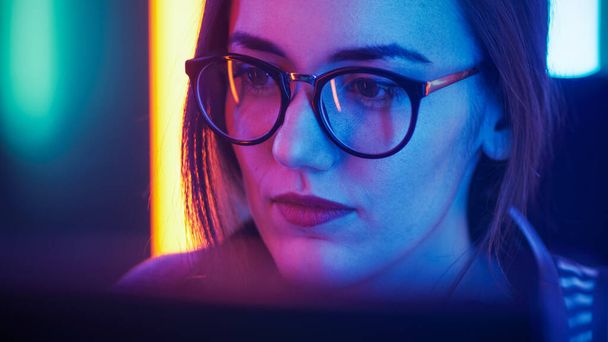 Portret pięknej młodej dziewczyny siedzącej przed komputerem, przeglądającej internet, grającej w gry online, streaming. Ładne dziewczyny noszące okulary w chłodnym pokoju Neon Lit Retro. - Zdjęcie, obraz