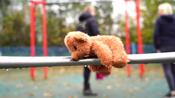 Lost Teddy bear lying on swing with rain drops in rainy day,Lonely or sad brown bear lied down alone with blurry kids and parent playing swing in the park background, International missing children's  - Footage, Video