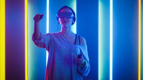 Beautiful Young Girl Wearing Virtual Reality Headset Using Joysticks Controllers. Creative Young Girl Does Concept Art with Augmented Reality. Playing Online Video Game. Neon Retro Lights Surround Her - 写真・画像