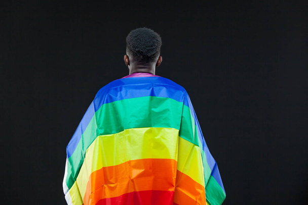 Back view of young african american man wrapped in a rainbow flag isolated on black background. Concept of The LGBT community, minority rights, protection of human rights - Foto, afbeelding