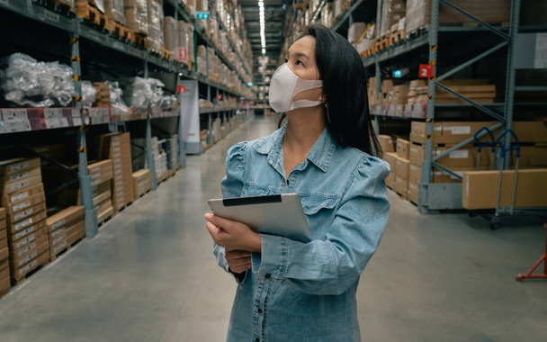 Empresa asiática mujer trabajadora usar máscara utilizando tableta digital comprobación de stock de productos en el almacén - Foto, Imagen