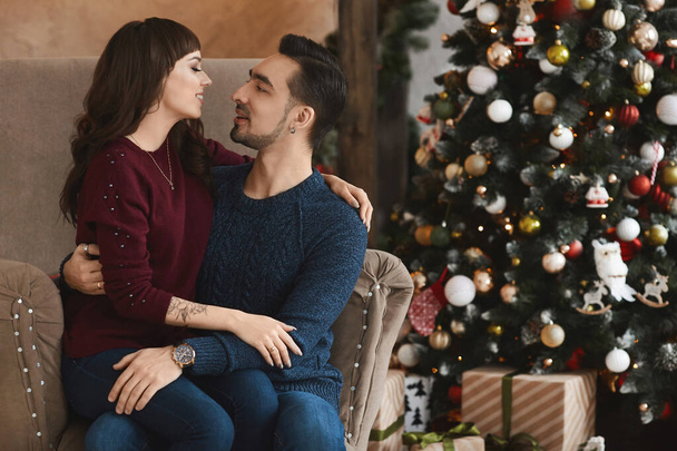Joven hombre guapo abraza a una chica morena en el sillón en una sala de estar en época de Navidad - Foto, Imagen