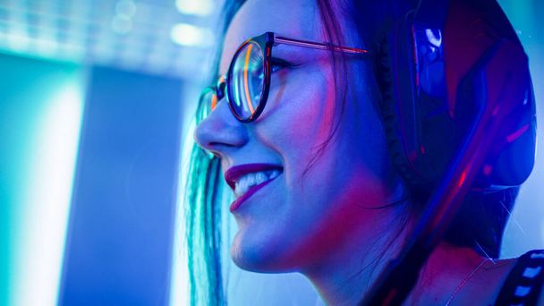 Low Angle Portrait Shot of the Beautiful Pro Gamer Girl Playing in Online Video Game, Cute Geek Girl in Glasses, talks with Team Players through Microphone. Neon Colored Room. e-Sport Cyber Games - Zdjęcie, obraz