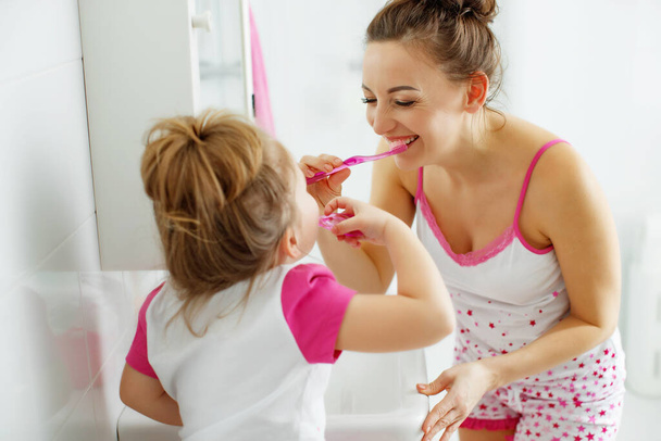 Мать и дочь чистят зубы зубными щетками в ванной комнате дома. Мама и девочка в домашней одежде. - Фото, изображение