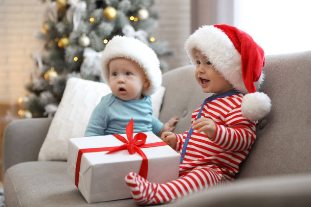 Cute children in Santa hats sitting on sofa at home. Christmas celebration - Фото, изображение