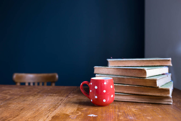 A stack of large, old, rag-bound books lie on a wooden table against a dark blue wall. Pen and notebook. Concept - back to school. Online training. Copy space. Select focus. Red cup with white polka dots with tea. - Photo, Image