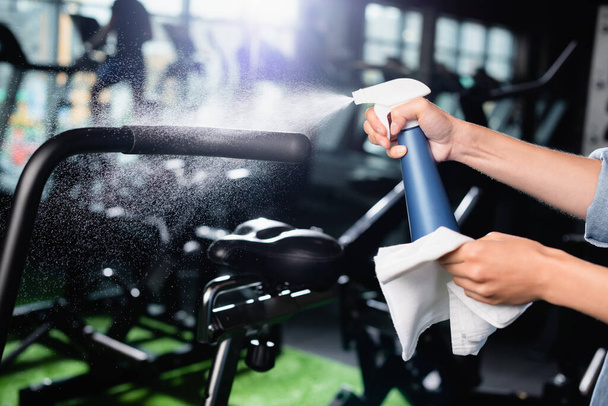 vista recortada del detergente de pulverización charwoman mientras se limpia la máquina de ejercicios en el gimnasio - Foto, imagen