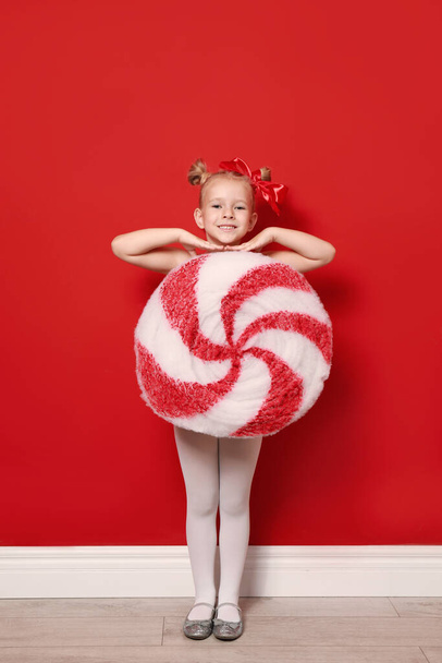 Cute little girl dressed as candy near red wall. Christmas suit - Фото, изображение