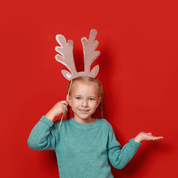 Cute little girl with reindeer antlers prop on red background. Christmas celebration - Foto, Imagem