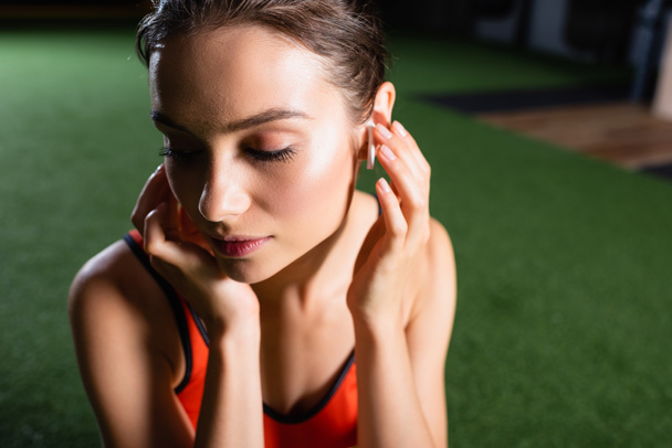 junge Sportlerin berührt drahtlosen Kopfhörer beim Musikhören im Fitnessstudio - Foto, Bild