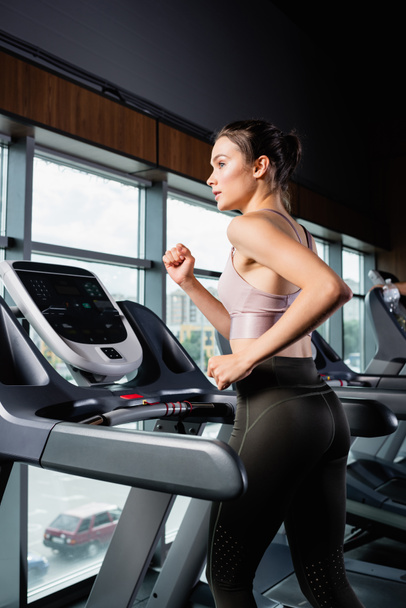 side view of sportswoman in leggings looking away while running on treadmill - Foto, Bild
