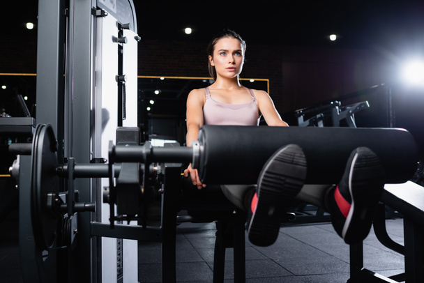 sport kobieta w biustonosz robi nogi wydłużenie ćwiczenia na maszynie treningowej na zamazanym pierwszym planie - Zdjęcie, obraz